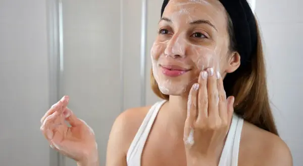Mujer con espuma en la cara, disfrutando de su rutina de limpieza facial en casa.