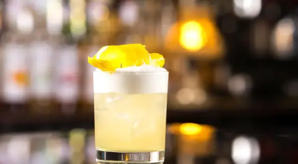 Klasyczny whisky sour przepis: orzeźwiający koktajl z pianką i skórką cytryny na tle rozmytego baru.