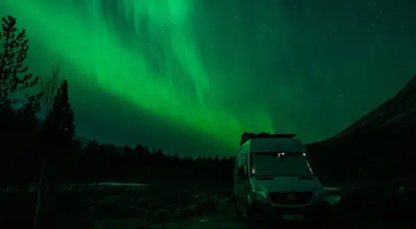 Ein Camper steht unter dem faszinierenden Licht der Aurora Borealis. Grüne Schleier tanzen am sternenklaren Himmel über dunklen Bäumen und einer Bergflanke.