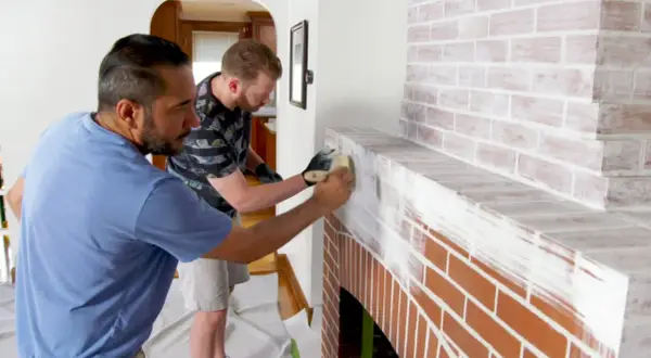 Two men are learning how to whitewash brick on a fireplace, applying white paint with brushes for a rustic look.