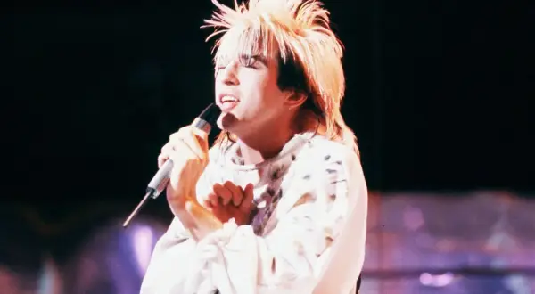 Limahl piosenkarz z charakterystyczną fryzurą śpiewa do mikrofonu na scenie.