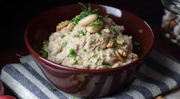 Aksamitna pasta z lubczyku, udekorowana fasolką, orzechami i ziołami, w bordowej misce na pasiastym obrusie.