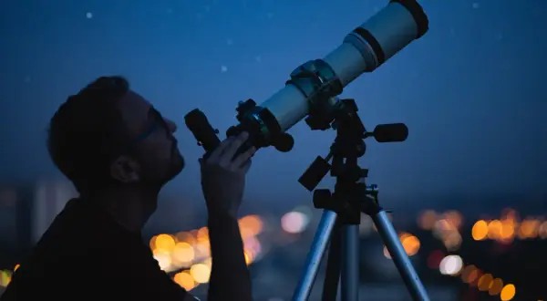 Mężczyzna obserwuje nocne niebo przez teleskop na tle rozświetlonego miasta.