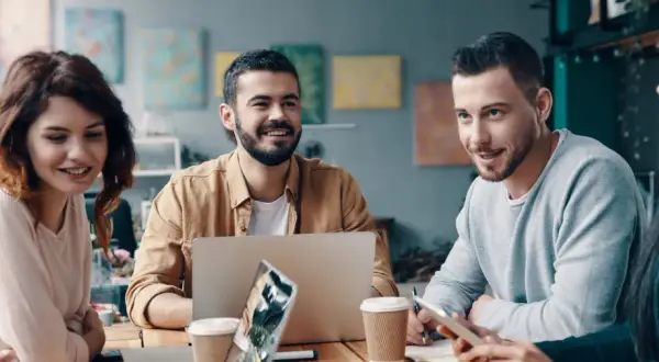 En grupp unga, leende människor diskuterar vid ett bord. En laptop och kaffekoppar antyder ett möte om utbildning, organisation och ledarskap.