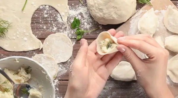 Ręce formują pieroga z ciasta na pierogi, wypełnionego farszem ziemniaczanym i koperkiem. Na drewnianym blacie widać mąkę, gotowe pierogi i miskę z farszem.