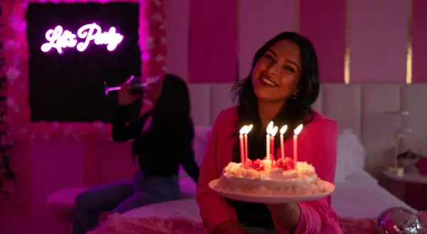 Urodziny Barbie w różowym pokoju. Uśmiechnięta dziewczyna trzyma tort ze świeczkami, a w tle koleżanka pije drinka.