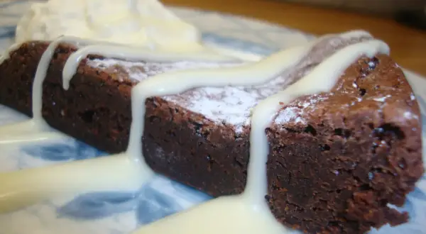 En bit mörk kladdkaka med pudersocker och vit chokladsås, serverad med vispad grädde.