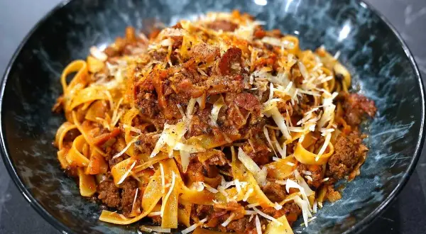 En skål med breda bandnudlar täckta av en mustig köttfärssås, toppad med riven ost och peppar. En härlig pasta ragu!