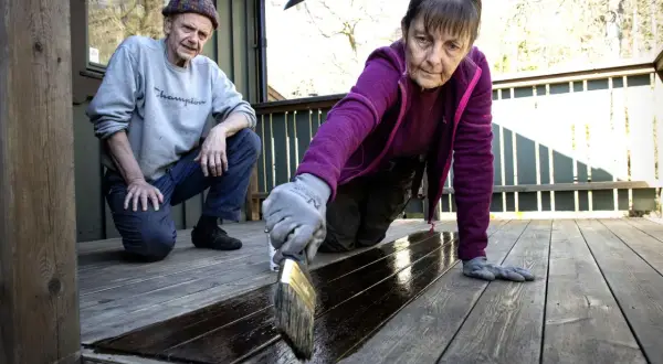 Kvinna penslar mörkt träskydd på altanen. Mannen tittar på. De vill lasera tryckimpregnerat trä för att skydda det.
