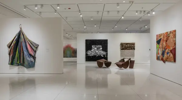 An art gallery features diverse works, including a colorful draped sculpture, a black and white textured piece, and a vibrant collage, reminiscent of the bold style of Pamela Joyner.
