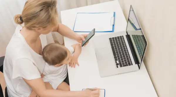 Kobieta z dzieckiem na kolanach sprawdza na telefonie, urlop macierzyński ile płatny. Obok laptop i notatnik.