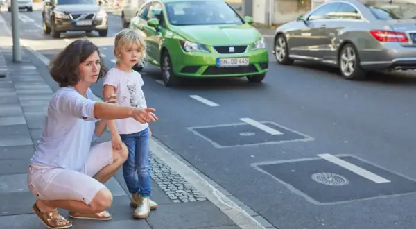 Mutter erklärt ihrer Tochter, dass sie auf dem Gehweg nicht in falsche Richtung fahren darf. Ein grünes Auto fährt auf der Straße.