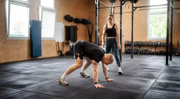 Mężczyzna wykonuje burpee pod okiem trenerki w siłowni.