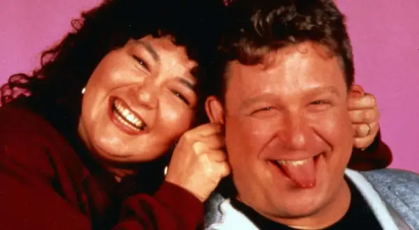 Roseanne und Dan, die Hauptfiguren der besetzung von Roseanne, lachen und Grimassen vor einem rosa Hintergrund.