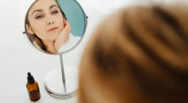 Kobieta w lustrze zastanawia się, czy jest "pick me girl", dotykając policzka. Obok buteleczka z serum.
