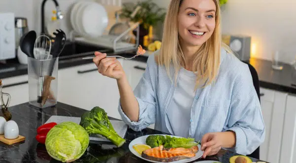 Femme souriante dégustant un menu équilibré pour perdre du poids : saumon, brocolis, avocat, noix et légumes frais.