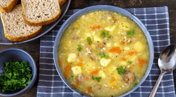 Gęsta zupa grochowa z ziemniakami, marchewką i kiełbasą. Obok kromki chleba i miseczka natki pietruszki – idealne dodatki do zupy grochowej.