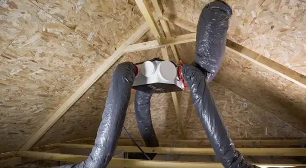 Installation d'une VMC dans un grenier. Le caisson blanc est suspendu, relié à des gaines grises flexibles.
