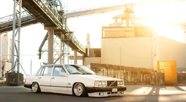 Vit Volvo 740 från 1990, sänkt och med kromade fälgar, parkerad vid en industrianläggning i solnedgången.
