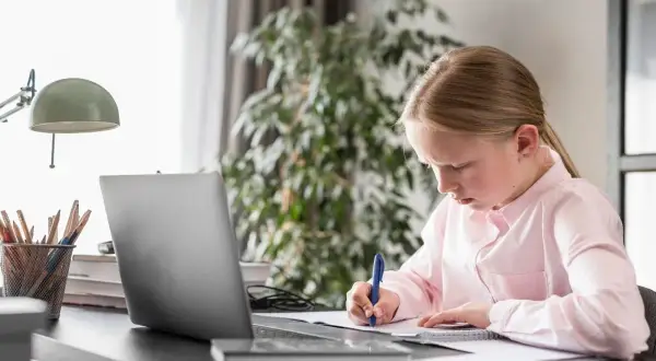 Dziewczynka uczy się w domu, pisząc notatki. Zastanawia się, ile kosztuje nauczanie domowe, analizując materiały na laptopie.