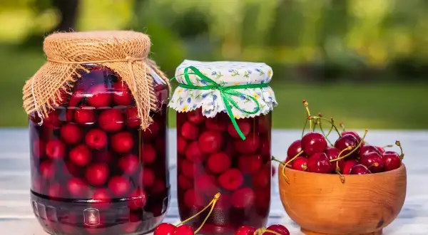 Dwa słoiki domowego kompotu z wiśni, obok miseczka świeżych owoców.