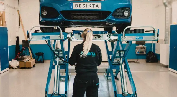 En bil lyfts upp på en lyft för sista besiktningsdag. En mekaniker med flätat hår står framför bilen.