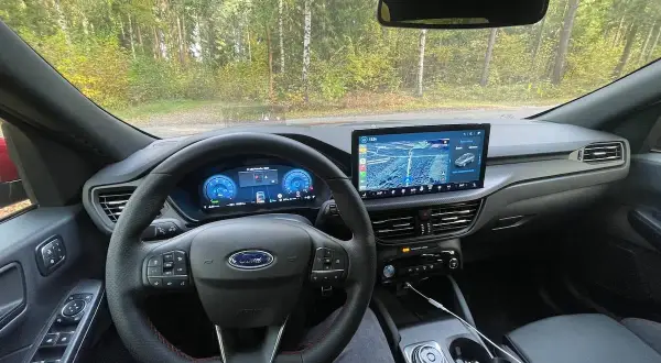Wnętrze Ford Kuga z nowoczesnym ekranem multimedialnym i cyfrowymi zegarami. Kierownica z czerwonymi przeszyciami i przyciskami.