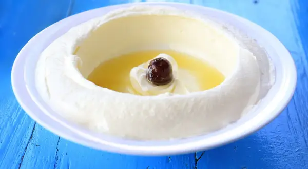 Un plato blanco con labneh que es un queso cremoso, con aceite de oliva y una aceituna negra en el centro, sobre una mesa azul.
