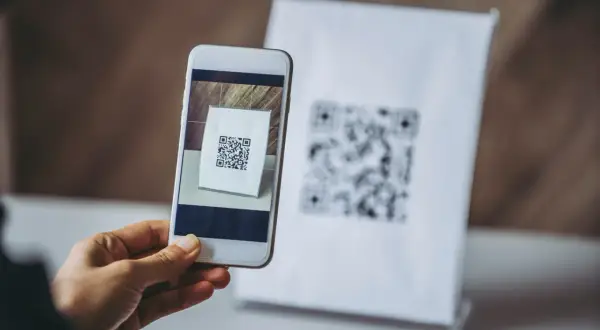 Dłoń trzyma smartfon skanujący kod QR na białej kartce.