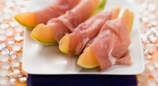 Saftige Honigmelone mit zartem Schinken umwickelt, serviert auf einem weißen Teller. Ein süß-salziger Genuss!
