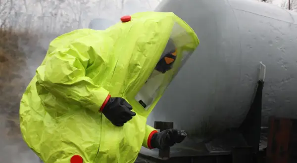 Person in a bright yellow hazmat suit with a clear visor, standing next to a large grey tank.