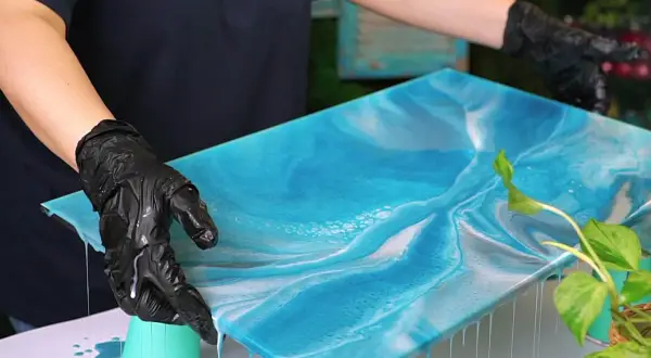 Mani guantate controllano un'opera d'arte in resina epossidica blu e bianca, simile al mare. Il tempo di indurimento della resina epossidica è cruciale per questo effetto.
