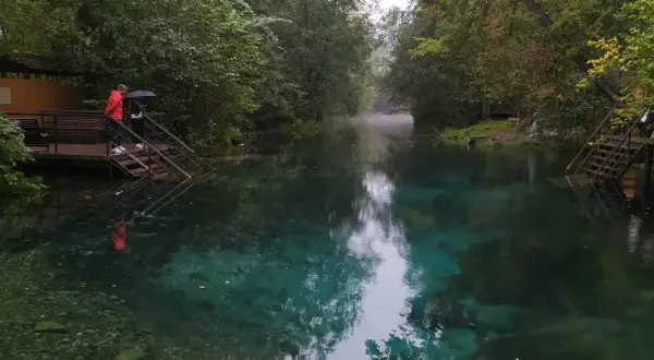 Turkusowe jezioro w Rosji, otoczone drzewami. Dwie osoby stoją na schodach prowadzących do wody, jedna z parasolem.