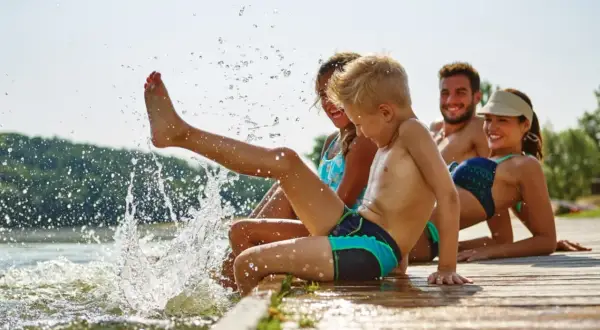 Familie genießt den Sommer am See. Kinder planschen im Wasser, Eltern entspannen sich. Guter uva schutz ist wichtig.