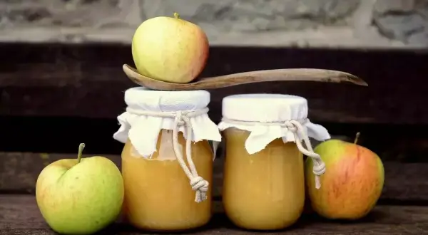 Deux pots de compote de pommes faits maison, décorés de tissus et de cordes, avec des pommes fraîches et une cuillère en bois.