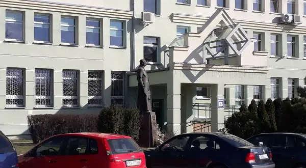 Pomnik przed budynkiem z oknami i kratami. Obok czerwony samochód, a dalej ciemny. KN rejestracja na tablicy.