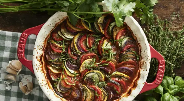 Vacker ratatouille i en röd ugnsform, med skivade grönsaker som tomat, zucchini och aubergine. Perfekt för ett ratatouille recept.
