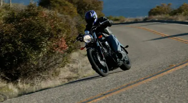 Motociclista inclinado en una curva, demostrando el **efecto giroscópico** de las ruedas al girar. El mar y la vegetación bordean la carretera.