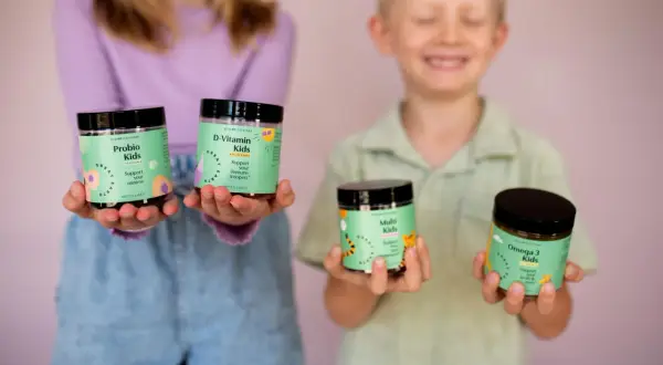 Två barn håller upp burkar med vitaminer. En flicka håller Probio Kids och D-Vitamin Kids, medan en pojke håller Multi Kids och Omega 3 Kids. Perfekt för vitamin barn!