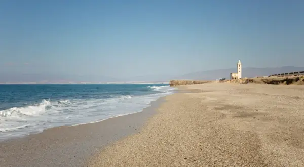 La almadraba de Monteleva, una torre blanca se alza en la playa, con el mar azul y las montañas al fondo.