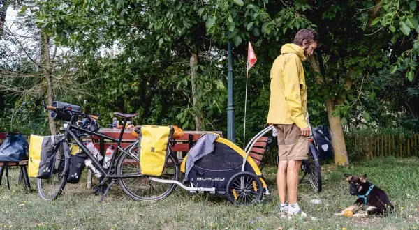Un homme en jaune se tient près d'un vélo chargé, avec une remorque et un jeune chien joueur. Un chien Canada prêt pour l'aventure.