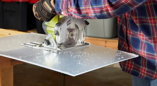 A person in a plaid shirt and work gloves uses a circular saw to cut a sheet of plexiglass, demonstrating how to cut plexiglass cleanly.
