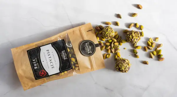 Opakowanie pistacji i trzy praliny z pistacjami rozsypane na marmurowym blacie. Warto sprawdzić pistacje kcal przed spożyciem.