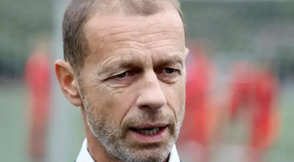 Aleksander Čeferin, prezes UEFA, w niebieskiej koszuli i granatowym swetrze, z rozmytym tłem piłkarzy.
