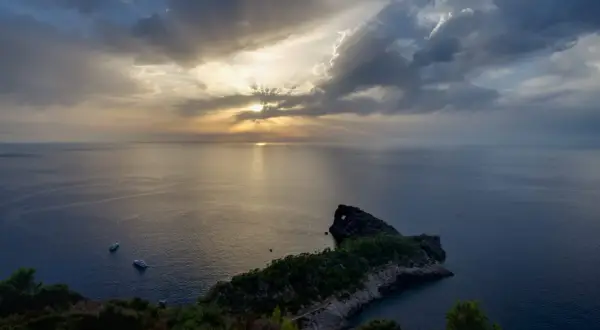 Atardecer en el mirador de Sa Foradada, con rayos de sol filtrándose entre nubes y dos barcos en el mar.