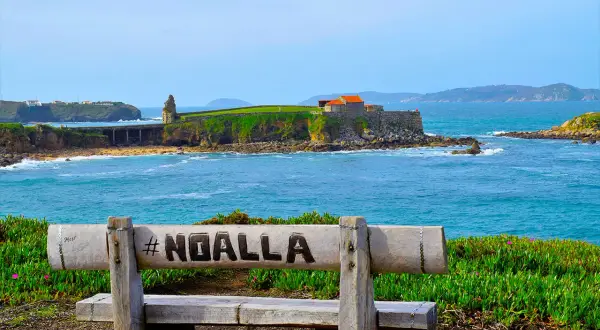 Banco de madera con #NOALLA grabado, frente al mar azul y un pueblo costero con edificios históricos.