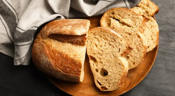 Krojony bochenek chleba i kromki na drewnianej desce, obok lnianej ściereczki.