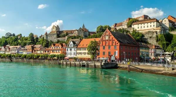 Meersburg Tourismus: malerische Altstadt mit bunten Häusern und Burg am Hang, überragt von grünen Hügeln, am glitzernden Bodensee.