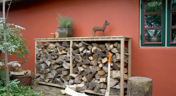 Praktische brennholz lagern ideen: Holzstapel in einem Holzregal vor einer roten Wand, daneben ein Baumstumpf und eine Axt.
