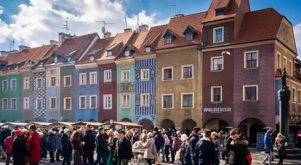Kolorowe kamienice na Starym Rynku w Poznaniu. Tłum ludzi spaceruje po bruku. Idealne miejsce, by poczuć atmosferę miasta, gdy już wiesz, jak dojechać z dworca głównego w Poznaniu na stary rynek.